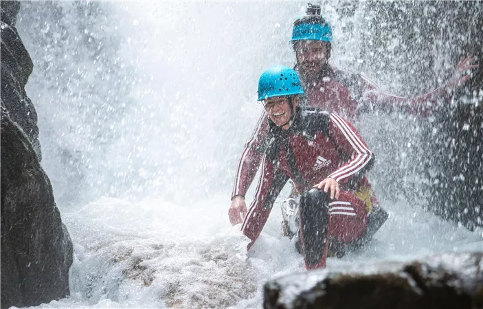Two cyclists on a narrow path in a green, hilly landscape with mountains in the background. The sky is blue with some clouds.