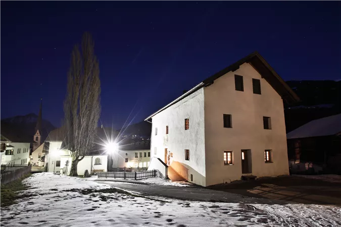 A quiet village at night with a snow-covered ground. The buildings are lit up and the sky is clear.