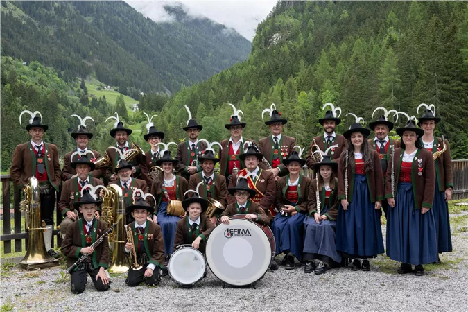 Eine Gruppe von Musikern in traditionellen Trachten steht in einer schönen Berglandschaft. Sie lächeln und posieren vor einem beeindruckenden grünen Hintergrund.