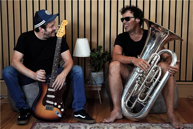 Zwei Musiker sitzen entspannt im Raum. Einer hält eine Gitarre, während der andere eine Tuba spielt.