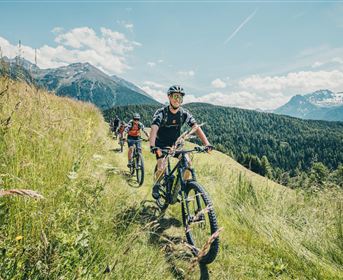 Zwei Radfahrer auf einem schmalen Pfad in einer grünen, hügeligen Landschaft mit Bergen im Hintergrund. Der Himmel ist blau mit einigen Wolken.