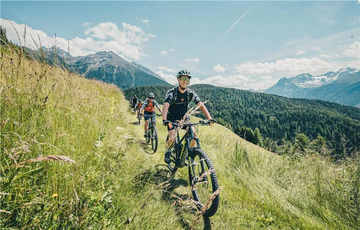 Two cyclists on a narrow path in a green, hilly landscape with mountains in the background. The sky is blue with some clouds.