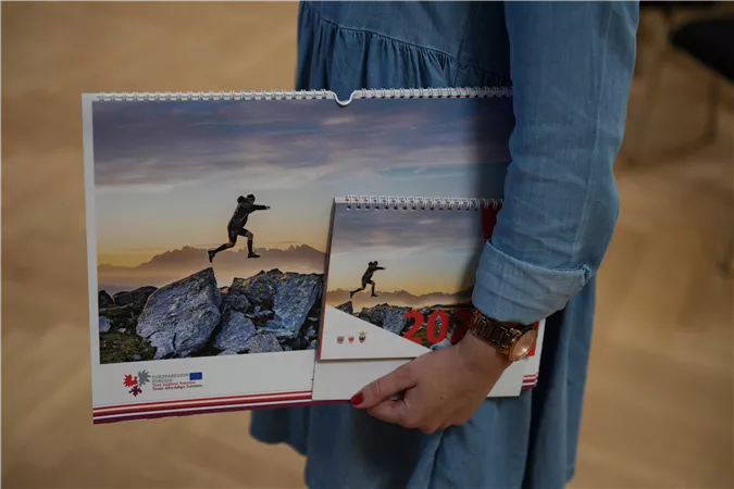 Eine Person hält einen großen und einen kleinen Kalender in der Hand. Der große Kalender zeigt einen Bergsteiger, der über Felsen springt, im Hintergrund sind Berge zu sehen.