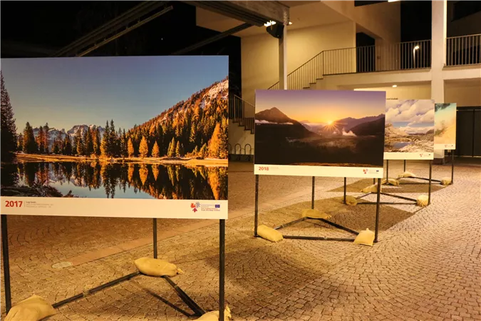 Eine Fotografienausstellung mit mehreren Bildern von Landschaften ist in einem modernen Raum aufgebaut. Die Bilder zeigen Naturansichten mit Bergen, Wäldern und Wasserflächen.