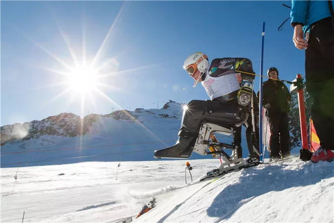 Ein Skater sitzt in einem speziellen Sitzschlitten und bereitet sich auf den Sprung. Im Hintergrund sind schneebedeckte Berge und ein sonniger Himmel zu sehen.