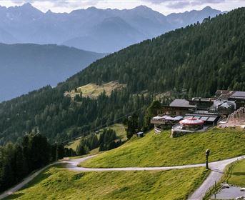 Eine malerische Berglandschaft mit grünen Wiesen und majestätischen Bergen im Hintergrund. Im Vordergrund liegt ein Wanderweg, der zu einer gemütlichen Hütte führt.