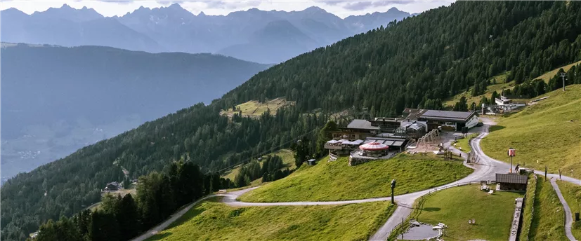 Eine malerische Berglandschaft mit grünen Wiesen und majestätischen Bergen im Hintergrund. Im Vordergrund liegt ein Wanderweg, der zu einer gemütlichen Hütte führt.