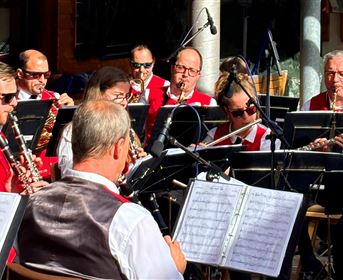 Eine Blaskapelle spielt in einem Freiluftkonzert. Die Musiker tragen rote Westen und konzentrieren sich auf die Aufführung.