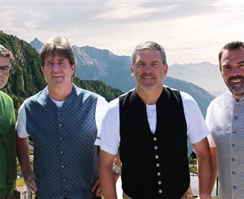 Vier Männer in traditioneller Tracht stehen vor einer schönen Bergkulisse. Im Hintergrund sind bewaldete Berge und ein klarer Himmel sichtbar.