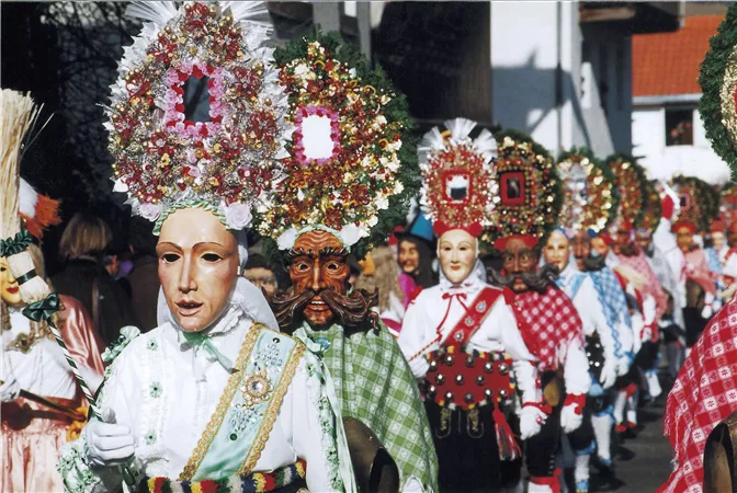 Eine bunte Parade mit Menschen in traditionellen Masken und festlichen Kostümen. Die Teilnehmer tragen blühende Kopfbedeckungen und feiern fröhlich.