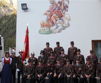 Eine Gruppe von Feuerwehrleuten in Uniform steht vor einem Feuerwehrfahrzeug und einem Gebäude mit einer Wandmalerei. Im Hintergrund sind Berge sichtbar.