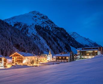 Eine malerische Winterlandschaft mit schneebedeckten Bergen und einem kleinen Dorf. Die Gebäude sind abends beleuchtet und verleihen der Szenerie eine gemütliche Atmosphäre.
