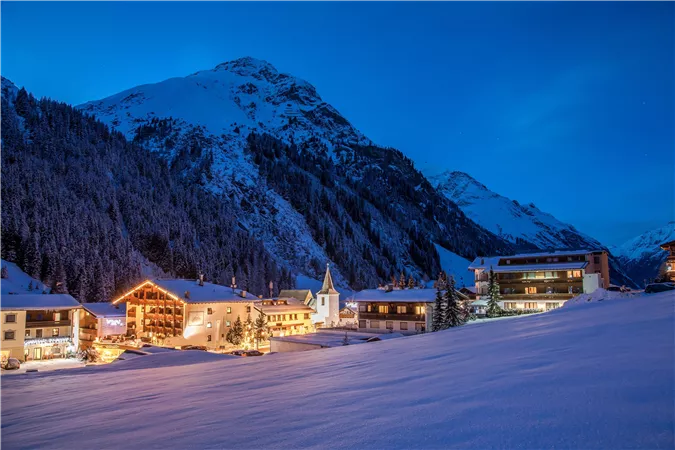 Eine malerische Winterlandschaft mit schneebedeckten Bergen und einem kleinen Dorf. Die Gebäude sind abends beleuchtet und verleihen der Szenerie eine gemütliche Atmosphäre.