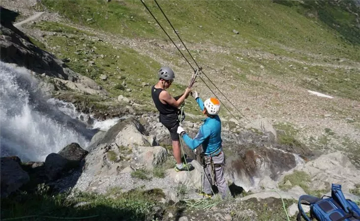 Zwei Kletterer arbeiten an einem Seil unterhalb eines Wasserfalls. Die Umgebung ist grün und bergig mit fließendem Wasser.