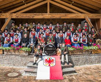 Eine Gruppe von Musikern und Traditionskleidung versammelt sich vor einem Holzgebäude. Im Vordergrund liegen eine Flagge und ein Preis.
