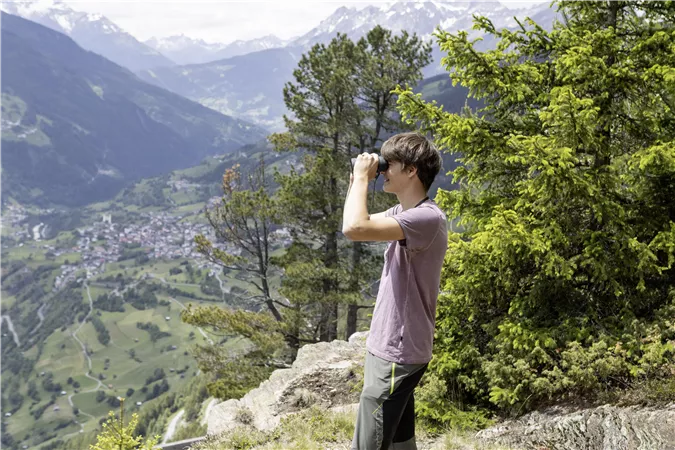 A young man stands on a mountain and observes the landscape with binoculars. The impressive mountains and the green valley are visible in the background.