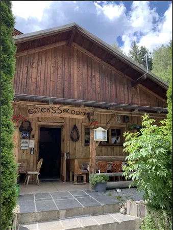 Ein rustikales Holzgebäude mit der Aufschrift "Eiter's Saloon". Der Eingang ist von Pflanzen und Stühlen umgeben und strahlt eine gemütliche Atmosphäre aus.