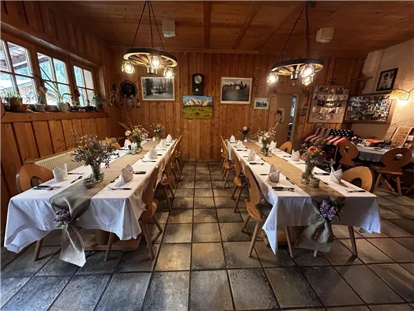 Ein rustikales Restaurant mit langen Tischreihen, dekoriert mit Blumen und weißen Tischdecken. Die Wände sind aus Holz und verleihen der Atmosphäre eine gemütliche Note.