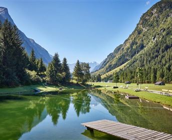 Ein ruhiger Bergsee umgeben von hohen Bäumen und grünen Wiesen. Die reflektierenden Berge im Hintergrund schaffen eine malerische Landschaft.