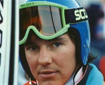 Ein Skifahrer mit einem konzentrierten Blick trägt eine bunte Ausrüstung und einen Helm. Im Hintergrund sind unscharfe Skiausrüstungen und Zuschauer zu sehen.