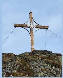 Ein großes Holzkreuz steht auf einem felsigen Hügel. Der Himmel ist klar und blau, was eine ruhige Atmosphäre schafft.