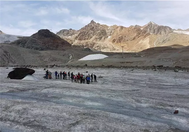 Eine Gruppe von Menschen steht auf einem Gletscher in den Bergen. Im Hintergrund sind majestätische Berggipfel und ein klarer Himmel zu sehen.