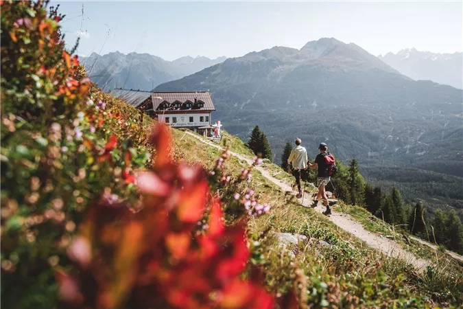 Zwei Wanderer gehen einen malerischen Pfad entlang, umgeben von bunten Blumen und Bergen. Im Hintergrund ist ein gemütliches Gebäude sichtbar.