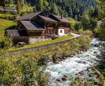 An idyllic wooden house by the shore of a clear brook, surrounded by green meadows and mountains. The tranquil landscape exudes rural beauty.