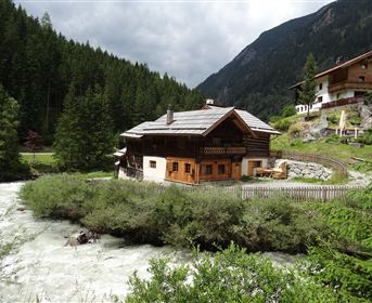 A guesthouse amidst green trees and a rushing river. In the background, mountains and additional buildings can be seen.