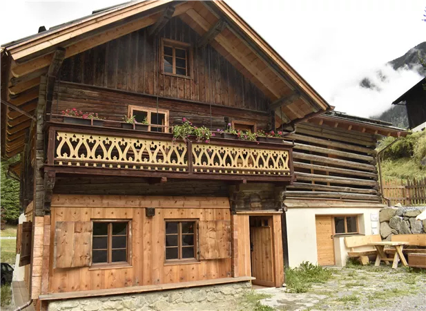 Ein traditionelles Holzhaus mit einem Balkon und Blumenkübeln. Umgeben von Natur und Bergen wirkt es einladend und gemütlich.