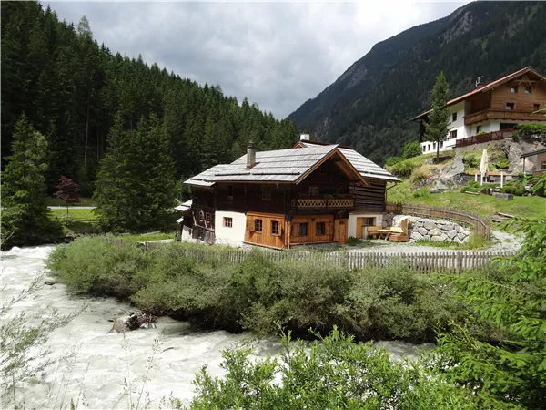 A traditional chalet-style house on the banks of a rushing river. In the background, high mountains and dense forests stretch out.