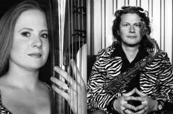 Two musicians in black and white. The woman is holding an instrument, and the man is sitting with his arms crossed.