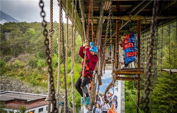 Two cyclists on a narrow path in a green, hilly landscape with mountains in the background. The sky is blue with some clouds.