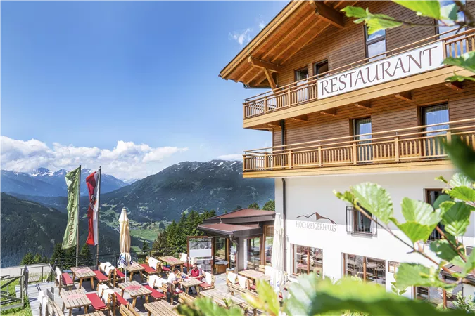 A cozy restaurant in the mountains with terrace seating and a view of nature. Clear blue skies and green surroundings create an inviting atmosphere.