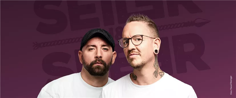 Zwei Männer stehen vor einem lila Hintergrund und schauen in die Kamera. Sie tragen weiße T-Shirts und haben jeweils verschiedene Stile, darunter Tattoos und eine Mütze.
