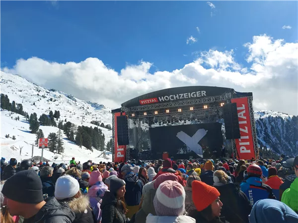 Eine große Menschenmenge steht vor einer Bühne in den Bergen, umgeben von Schnee. Der Himmel ist blau mit einigen Wolken.