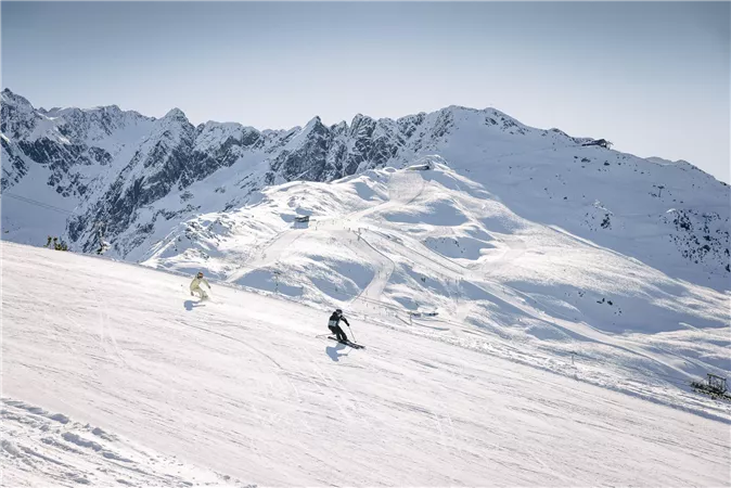 Zwei Skifahrer fahren eine verschneite Piste hinunter. Im Hintergrund sind majestätische Berge unter einem klaren Himmel zu sehen.