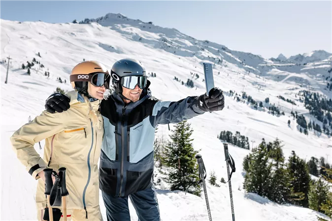 Ein Skipaaar posiert auf den schneebedeckten Pisten in den Bergen. Sie lächeln und machen ein Selfie vor der winterlichen Kulisse.