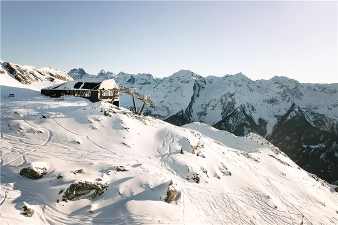Eine schneebedeckte Berglandschaft mit majestätischen Bergen im Hintergrund. Im Vordergrund steht eine Berghütte und Skispuren führen durch die glitzernde Schneedecke.
