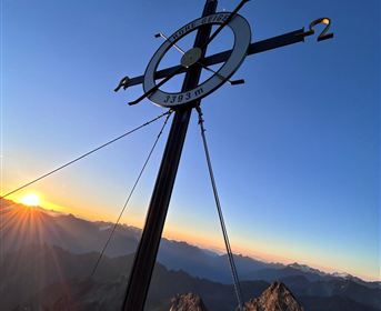 Ein Gipfelkreuz vor einem Sonnenuntergang über den Bergen. Die warme Lichtstimmung hebt die Schönheit der Landschaft hervor.