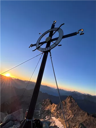 Ein Gipfelkreuz vor einem Sonnenuntergang über den Bergen. Die warme Lichtstimmung hebt die Schönheit der Landschaft hervor.