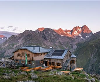 A mountain cabin surrounded by majestic mountains. In the background, the first rays of sunlight announce the day.