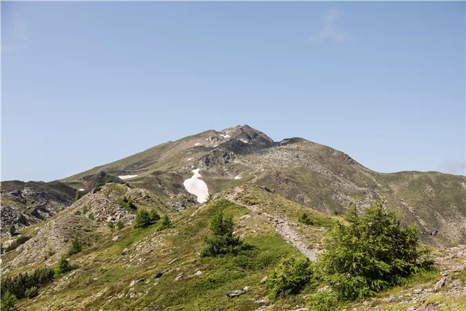 Ein Berg mit grünen Wiesen und einem kleinen Schneefeld. Der Himmel ist klar und blau.