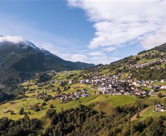A picturesque mountain landscape with a small village and green meadows. In the background, snow-capped mountains and a blue sky can be seen.