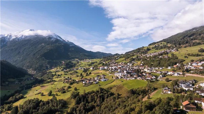 A picturesque mountain landscape with a small village and green meadows. In the background, snow-capped mountains and a blue sky can be seen.
