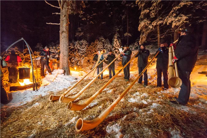 Eine Gruppe von Menschen spielt natürliche Hörner in einer verschneiten Umgebung. Im Hintergrund sind Feuer und Bäume zu sehen.