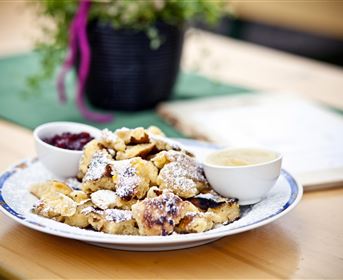 Ein Teller mit Kaiserschmarrn, bestäubt mit Puderzucker, liegt auf einem Tisch. Zwei Schalen mit Früchtekompott und Vanillesoße ergänzen das Gericht.