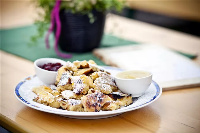 A plate of Kaiserschmarrn, dusted with powdered sugar, lies on a table. Two bowls of fruit compote and vanilla sauce complement the dish.