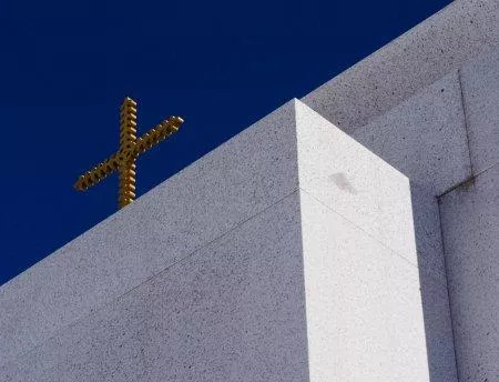Ein modernes Gebäude mit einer weißen Fassade und einem goldenen Kreuz oben. Der Himmel ist klar und blau.