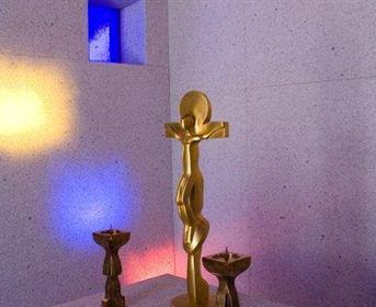 Eine goldene Skulptur steht im Raum, umgeben von zwei kleinen, dunklen Figuren. Bunte Lichtstrahlen fallen auf die Wände und schaffen eine ruhige Atmosphäre.